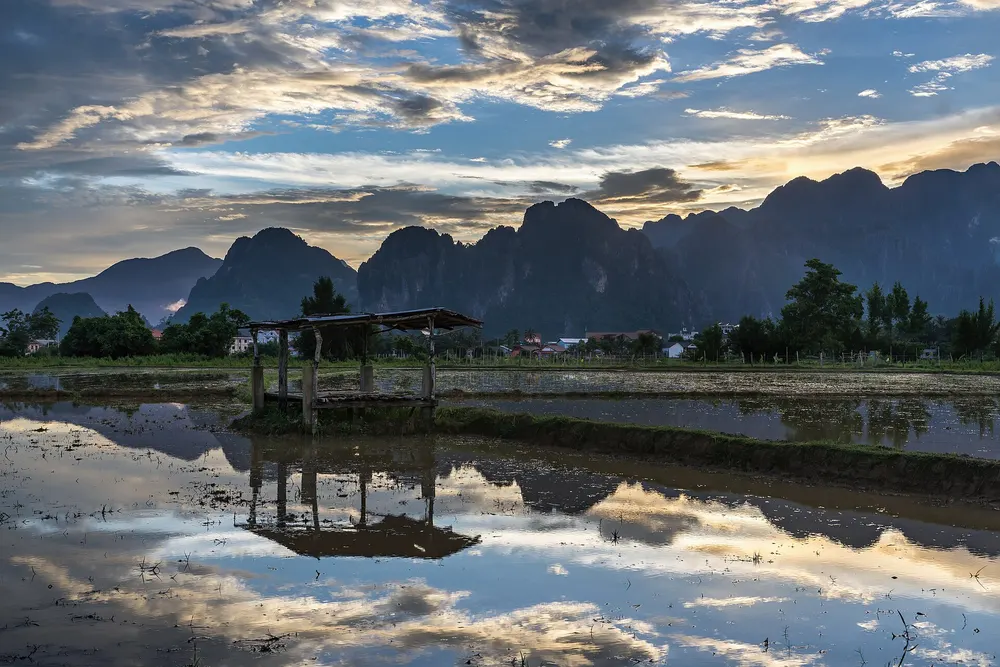 A wooden hut on a paddy field, its reflection in the water under a sunset sky